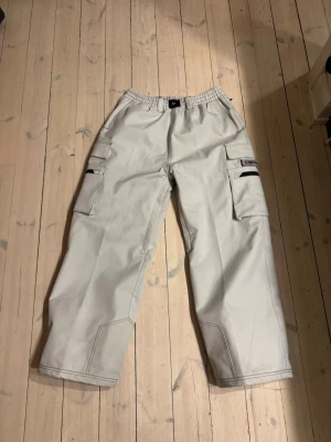 Beiga skidbyxor från Capeesh - Säljer ett par beiga skidbyxor från Capeesh med bred passform och elastisk midja. Byxorna har flera stora fickor med dragkedja och patchdetaljer, samt broderade svarta mönster på baksidan. Har en liten fläck på bakfickan som säkert går bort. Endast använda 1 gång 