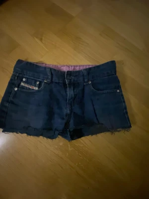 Lågmidjade mörkblå jeansshorts från G-Star - Säljer ett par mörkblå jeansshorts från G-Star med rå kant nertill och klassiska fem fickor. Shortsen har låg midja och silverfärgade nitar. Insidan är fodrad med rosa-rutigt tyg för en extra cool detalj.