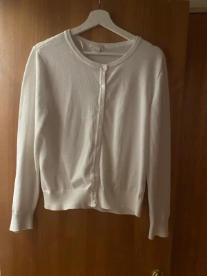 Vit kofta från H&M BASIC, storlek L - En enkel och stilren vit kofta från H&M BASIC i storlek L. Koftan har rund halsringning, långa ärmar och knäpps med små vita knappar framtill. Perfekt att slänga över en topp för en clean look.