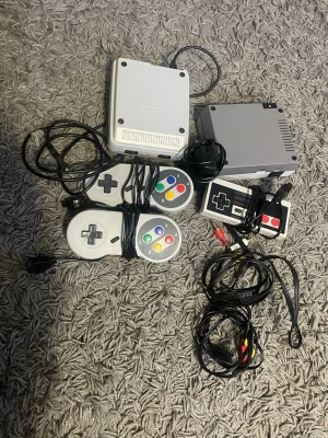 Repro spelkonsolpaket (NES & SNES-kloner)  EJ ORIGINAL  - EJ ORIGINAL utan kloner men fungerar bra och man kan uppleva många klassiska spel som super Mario osv. Paket med två repro spelkonsoler (NES- och SNES-kloner) inklusive tre kontroller och nödvändiga kablar. Perfekt för klassiska spelupplevelser. Konsoler och tillbehör i gott skick, endast mindre bruksspår synliga. Strömadapter och AV-kablar medföljer.