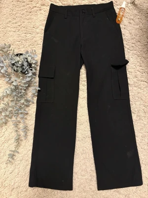 Svarta kostymbyxor från Kappahl stl 146 - Säljer ett par svarta cargobyxor från Kappahl i storlek 146. Byxorna har raka ben, praktiska sidofickor med lock och bälteshällor i midjan. Perfekta för en avslappnad och trendig look.