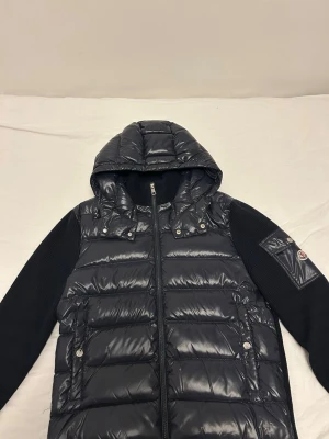 Marinblå Moncler cardigan   - Säljer en marinblå pufferjacka från Moncler med glansig finish och huva. Jackan har stickade ärmar och två fickor framtill med knappstängning. Klassisk Moncler-logga på ena ärmen och dragkedja framtill. Perfekt för kyliga dagar. I mycket bra skick, köpt från merchsweden och har endast använt den någon enstaka gång.