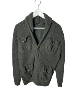 Mörkgrön stickad kofta från Tiger of sweden - Säljer en mörkgrön, ribbstickad kofta från Tiger med fyra fickor framtill och snygga träknappar. Koftan har bred krage och långa ärmar, perfekt för lager på lager under kyliga dagar.