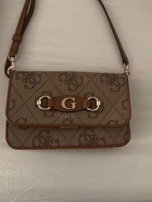 Beige axelväska från Guess - Säljer en beige axelväska från Guess med bruna detaljer och logomönster över hela väskan. Väskan har ett stort G i metall framtill och justerbar axelrem. Perfekt storlek för det viktigaste och snygg design med metallspännen.