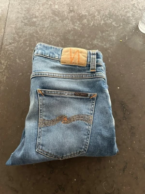 Ljusblå jeans från Nudie Jeans - Säljer ett par ljusblå jeans från Nudie Jeans med orange sömmar. Jeansen har en slim passform och är tillverkade i mjukt denimtyg. Det är också uppsydda i botten. W29 och Längd var 32! Riktigt sköna och perfekta inför våren! 