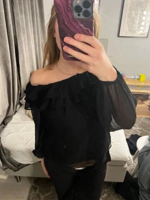 Svart offshoulder volangblus Gina Tricot - Säljer en svart offshoulder blus från Gina Tricot med volangdetalj längs ringningen och tunna, transparenta långärmar. Blusen har en luftig och flowy passform som ger en snygg siluett. Perfekt för dig som gillar trendiga detaljer och stilren look.