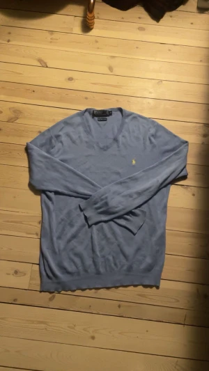 Blå v-ringad tröja från Polo Ralph Lauren - Snygg blå v-ringad tröja från Polo Ralph Lauren med den klassiska gula loggan broderad på bröstet. Tröjan har lång ärm och är tillverkad i mjukt material som passar perfekt för lager-på-lager-stil. Storlek M. Hör av dig vid frågor!                                                             Byten kan vara intressant!