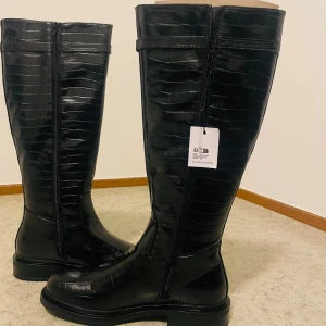 Svarta stövlar från Anna Field - Säljer ett par svarta höga stövlar från Anna Field med croco-mönstrad yta och dragkedja längs sidan. Stövlarna har platt sula och rund tå, tillverkade i syntetmaterial som ger en glansig finish. Perfekt för dig som gillar stilrena och trendiga boots. Den är helt ny