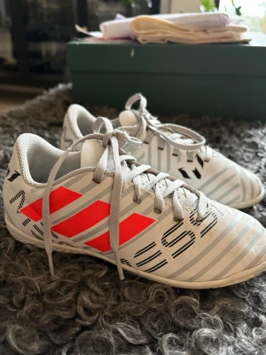 Adidas fotbollsskor - Säljer ett par Adidas inneskor i vitt med neon orange, ljus gråa och svart detaljer. Skorna har snörning och ett randigt mönster över hela ovandelen. Materialet är syntet med mesh vid öppningen för extra ventilation. Snygg, sportig design med platt sula och rund tå.