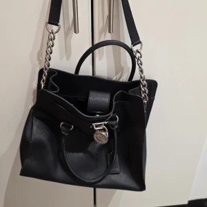 Svart handväska Michael Kors skinn - Snygg svart handväska från Michael Kors i skinn med silverfärgade kedjedetaljer och MK-hänglås framtill. Väskan har både handtag och axelrem, vilket gör den enkel att bära. Stilren design med metallbeslag och praktisk storlek för vardagsgrejer.