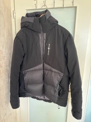 Svart dunjacka Sail Racing XL - Säljer en svart Sail Racing Dumont Down Jacket i storlek XL. Jackan har en schysst puffer-look med huva, flera dragkedjor och logga på bröstet. Fylld med dun och har paneler i olika material för extra stilpoäng. Perfekt för kalla dagar när du vill hålla värmen och se cool ut.
