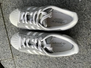 Adidas Superstar grå/vita sneakers - Adidas Superstar sneakers i grått/silver och vitt med klassiska tre ränder på sidan. Skorna har snörning och rund tå. Knappt använda. Perfekt för dig som gillar retrostil och streetwear.