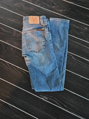 Blå Nudie Jeans Skinny W25 L32 - Säljer ett par snygga blå jeans från Nudie Jeans i modellen skinny. De har klassisk femficksdesign, orangea sömmar och Nudie-logga på bakfickan. Jeansen är i stretchigt denim och har en smal passform som sitter tajt längs benen.