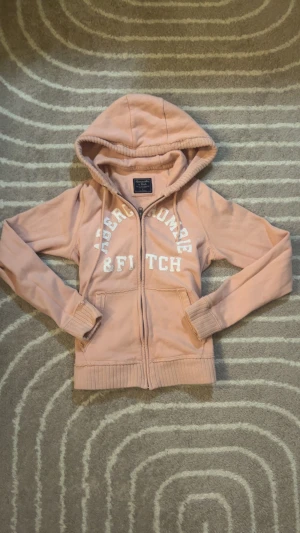 Rosa hoodie från Abercrombie & Fitch - Snygg ljusrosa hoodie från Abercrombie & Fitch med vit logotyp framtill. Tröjan har huva med snörning, dragkedja och ribbade muddar vid ärmslut och nederkant. Perfekt för chill dagar och har två fickor framtill.