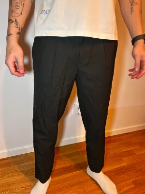 Svarta kostymbyxor med tapered fit - Snygga svarta kostymbyxor med tapered passform och croppad längd. Byxorna har pressveck framtill, sidfickor och bakfickor med knapp. Perfekt för dig som gillar stilrena och moderna byxor med lite smalare ben.
