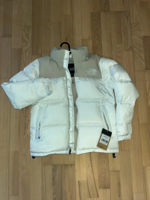 Vit och beige dunjacka The North Face - Säljer en riktigt snygg pufferjacka från The North Face i vitt och beige. Jackan har hög krage, två sidofickor med dragkedja och klassisk logga på bröstet. Perfekt för kalla dagar och har en clean, modern look.
