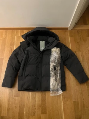Canada Goose Wyndham  - Säljer en svart dunjacka från Canada Goose i storlek L. Jackan har en pufferdesign med huva och medföljande pälsdetalj. Klassisk logga på ärmen, dragkedja och knappar framtill. Perfekt för kalla vinterdagar och riktigt snygg stil.