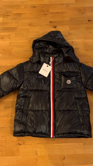 Svart Moncler pufferjacka med huva - Svart pufferjacka från Moncler med glansig finish, huva och ikonisk logga på bröstfickan. Jackan har en röd och vit rand längs dragkedjan, flera fickor och quiltad design. Insidan har ett serietryck och etiketter. Perfekt för kalla dagar.