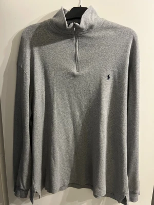 Grå half zip tröja från Polo Ralph Lauren XL - Snygg grå långärmad tröja från Polo Ralph Lauren med half zip och klassisk krage. Tröjan är i 100% bomull och har den ikoniska Polo-loggan broderad på bröstet. Perfekt för dig som gillar stilrena och bekväma plagg.