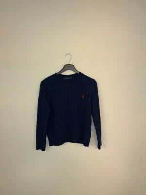 Marinblå stickad tröja Polo Ralph Lauren - Snygg marinblå stickad tröja från Polo Ralph Lauren i storlek barn L Tröjan har kabelstickat mönster och en röd broderad logga på bröstet. Tröjan passar både till vardag och när du ska klä upp! 