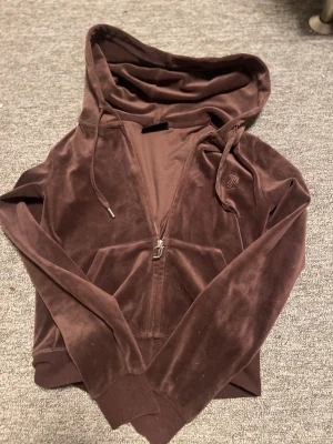Brun velour hoodie från Juicy Couture - Säljer en brun hoodie från Juicy Couture i mjuk velour med huva och snörning. Tröjan har dragkedja framtill, långa ärmar och fickor. JC-logga broderad på bröstet och snygga detaljer på dragkedjan. Perfekt för chill dagar.