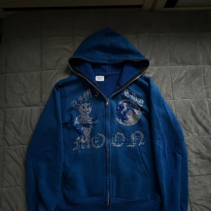 Blå rinestone hoodie med tryck - Blå hoodie med dragkedja och huva. Framsidan har ett coolt tryck med texten 'Aim for the moon', en figur och en måne i strass. Säljer denna zip hoodien då jag inte använder den mer. Jag säljer den för 500kr. Den har inga skador. 