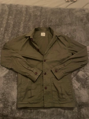 Olivgrön overshirt från John Henric - Snygg olivgrön overshirt från John Henric med klassisk krage och stora fickor framtill. Jackan har mörkröda knappar och är tillverkad i bomull, perfekt för lager-på-lager. Långärmad och med en clean, stilren look som funkar till många outfits.