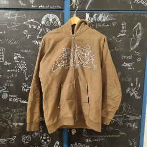 Brun hoodie med graffiti från HUF - Lätt använd HUF hoodie, den krympte i tvätten :( Köpt på Highrollers i Dublin ca 1 år sen för £145   100% cotton fleece (400GSM) heavyweight full zip hoodie 3D embroidered artwork at center chest Kangaroo pockets with metal rivets Two-way metal zipper front closure 2x1 rib-knit at cuffs and hem HUF woven label at interior neck