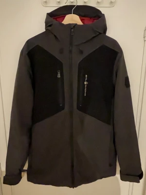 Svart Sail Racing jacka - Svart och mörkgrå vindjacka från Sail Racing med röd insida, huva och dragkedja framtill. Jackan har två bröstfickor med dragkedja och Sail Racing-logga på bröstet. Materialet är vind- och vattentätt, andas och har Primaloft-foder. Perfekt för blåsiga dagar.