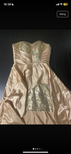 Champagnefärgad långklänning med paljetter - Champagnefärgad långklänning med glittrig överdel och paljetter, samt dekorativa stenar vid midjan. Klänningen är axelbandslös med snörning i ryggen och har en glittrig detalj på kjolen. Materialet är satinliknande och ger ett lyxigt intryck.