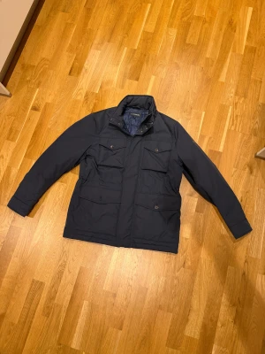 Mörkblå fieldjacket från J.Lindberg - Snygg mörkblå fieldjacket från J.Lindberg med fyra stora fickor framtill, dragkedja och knappar. Jackan har hög krage och långärmade ärmar, perfekt för kyliga dagar. Materialet är slitstark polyester och passformen är normal. Nypris ~4000