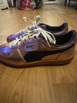 Puma Palermo sneakers metallic rosa - Säljer ett par Puma Palermo sneakers i metallic rosa med svarta detaljer och vit snörning. Skorna har en platt gummisula och rund tå. Materialet är syntet med en glansig finish som ger en riktigt cool vibe. Perfekt för dig som vill sticka ut med din stil.