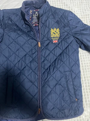 Marinblå quiltad jacka från Morris - Snygg marinblå quiltad jacka från Morris med gul broderad logga och brittisk flagga på bröstet. Jackan har dragkedja, ståkrage i manchester och praktiska fickor. Perfekt för dig som gillar klassisk stil med en modern twist.