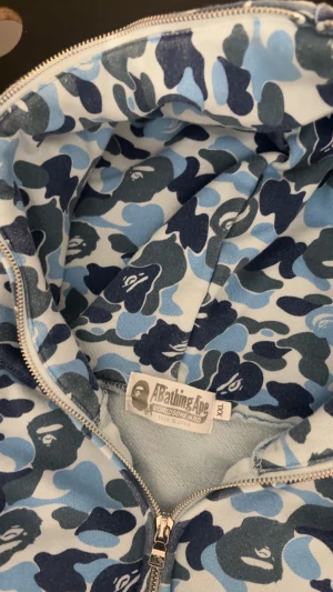 BAPE Shark Hoodie blå camo XXL - Säljer en ikonisk BAPE Shark Hoodie i blått camo-mönster med dragkedja hela vägen och huva med broderad hajdesign. Tröjan har ribbade muddar, fickor fram och detaljer i orange på sidan av huvan. Perfekt för dig som vill sticka ut med streetwear.