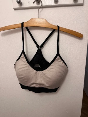 Beige och svart sport bh - Säljer en beige och svart sport bh  från soc med smala axelband som korsas i ryggen. Toppen elastiskt band under bysten och meshdetalj i ryggen. 