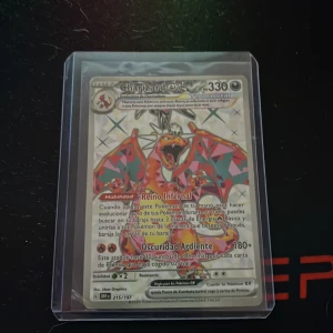 Charizard Pokémonkort 215/197 - Säljer ett Charizard Pokémonkort på spanska, nummer 215/197 från Scarlet & Violet-serien. Kortet har holografisk yta och motivet visar Charizard med färgglada vingar. Kortet är inplastat i ett skyddsfodral.