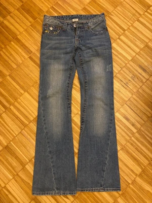 True Religion bootcut jeans blå, stl 26 - Säljer ett par klassiska blå jeans från True Religion med bootcut passform och snygga gula kontrastsömmar på bakfickorna. Jeansen är tillverkade i 100% bomull och har låg midja samt detaljerade fickor med knappar. Perfekt för dig som gillar ikoniska denim-vibes.