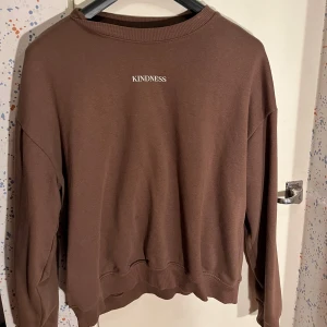 Brun sweatshirt - Säljer en brun sweatshirt med texten KINDNESS tryckt framtill. Tröjan har rund hals, långa ärmar och ribbade muddar vid ärmslut och nederkant. Storlek S.