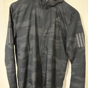 Svart camo vindjacka från Adidas  - Svart och grå vindjacka från Adidas i camomönster. Jackan har huva, hel dragkedja framtill och reflekterande ränder på ärmarna. Storlek S men passar M