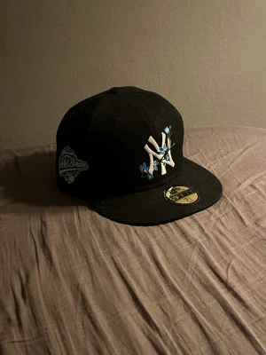 NY keps - Snygg svart New Era 59FIFTY keps med NY Yankees-logga framtill, dekorerad med broderade blå och vita blommor. På sidan finns ett World Series-märke i blått och vitt. Platt skärm och klassisk passform.