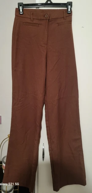 Bruna kostymbyxor med wide fit - Snygga bruna kostymbyxor med breda ben och klassisk midja. Byxorna har raka fickor framtill och stängs med knapp och dragkedja. Perfekta för en stilren look och passar dig som gillar lite vidare passform.