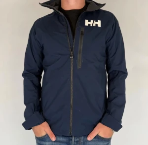 Marinblå vindjacka från Helly Hansen - Snygg marinblå vindjacka från Helly Hansen med vit logga på bröstet och tryck längs ena ärmen. Jackan har hel dragkedja framtill, en bröstficka med dragkedja och stårkrage. Perfekt för dig som gillar sportig och clean stil.