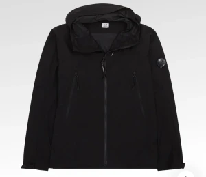 Svart vindjacka från C.P. Company - Svart vindjacka från C.P. Company med huva och ikonisk linsdetalj på ärmen. Jackan har två snedställda fickor med dragkedja, justerbar huva med snörning och hel dragkedja framtill. Perfekt för dig som gillar stilren och funktionell design. Nypris 4000kr