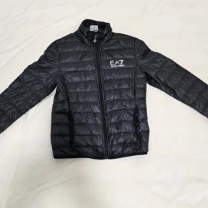 Svart dunjacka från EA7 Emporio Armani - Snygg svart dunjacka från EA7 Emporio Armani med quiltad design och hög krage. Jackan har dragkedja framtill, två sidofickor och logga på bröstet. Perfekt för dig som vill ha en stilren och lätt jacka med sportig känsla.