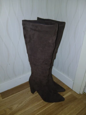 Bruna höga stövlar med klack - Säljer ett par snygga bruna stövlar i mocka med hög klack och spetsig tå. Stövlarna går över knäet och har en stilren, slät design som ger en cool och trendig look. Perfekta för dig som vill sticka ut med ett par statement boots.