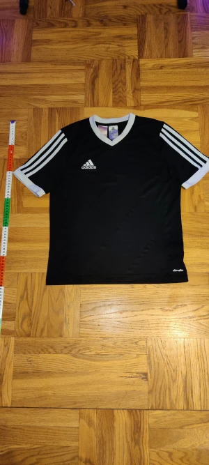 Svart Adidas t-shirt med vita detaljer - Svart Adidas t-shirt med vita ränder på axlarna och vita muddar vid ärmslut och krage. Klassisk v-ringning och broderad Adidas-logga på bröstet. Tillverkad i lätt och funktionell climalite-polyester, perfekt för sport och träning.