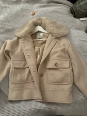 Beige kappa med fuskpäls från Neo Noir - Snygg beige kappa från Neo Noir med stor fluffig fuskpäls-krage, två stora fickor framtill och knappar. Jackan har klassisk rak passform och är perfekt för kyligare dagar. Materialet känns mjukt och lyxigt.