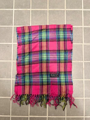 Rutig halsduk från Burberrys i ull - Klassisk rutig halsduk från Burberrys i 100% ull. Halsduken har fransar i ändarna och den ikoniska Burberrys-etiketten. Perfekt accessoar för att lyfta din outfit. Anledningen att de stör ”burberrys” är för att de är en äldre modell.