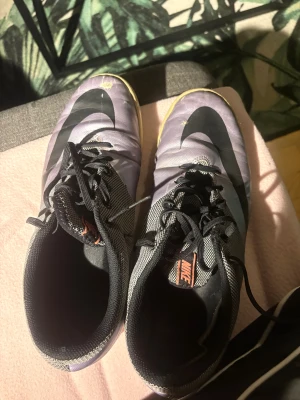 Lila/grå futsalskor LIMITED EDITION  - Säljer ett par gråa Nike fotbollsskor med svart swoosh och snörning. Skorna säljs inte längre i butik. Perfekta skor för dig som vill spela fotboll inomhus eller bara idrotta inomhus, och är även lätta och smidiga. Obs! Skorna är lite slitna på ovansidan samt har ett litet hål i sulan. Men fungerar att använda utan problem. Strl 45.5 men passar även 44. PRIS KAN DISKUTERAS!