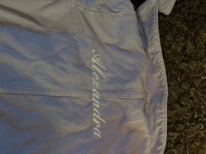 Ljuslila jeansjacka från Arket  - Säljer en snygg ljuslila jeansjacka från Arket med klassisk krage, tryckknappar och broderat namn 'Alexandra' på ryggen. Jackan har raka fickor framtill och är tillverkad i bomull. Perfekt för dig som vill sticka ut med en personlig touch.